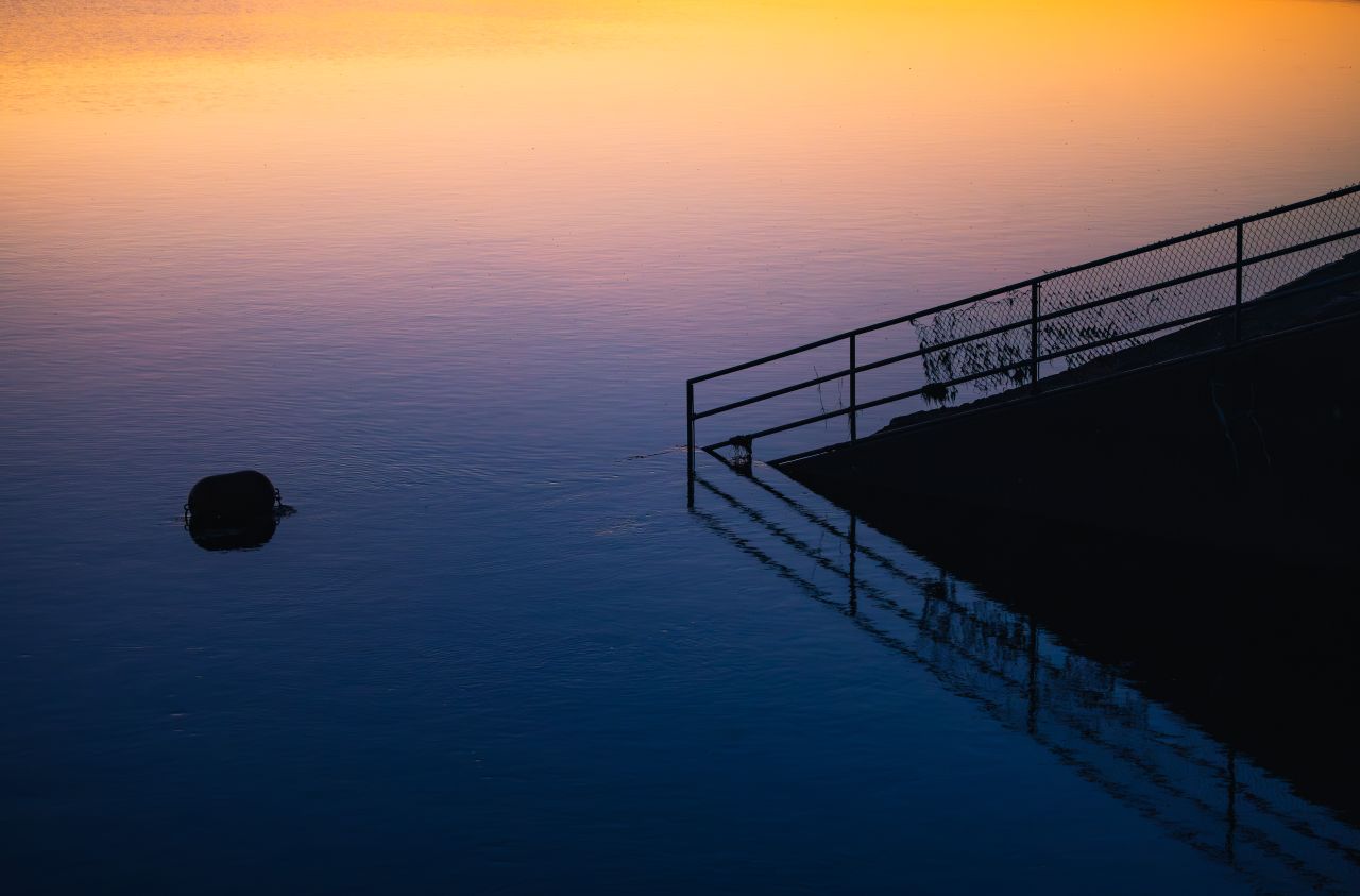 A serene sunset scene over calm waters with reflected hues and a silhouette of a fence.